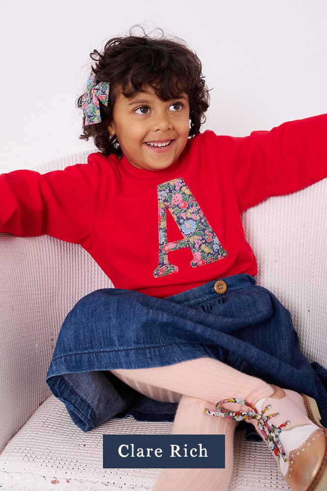 A 3 year old girl wearing a red personalised t-shirt with a liberty floral letter A appliquéd on the front. 