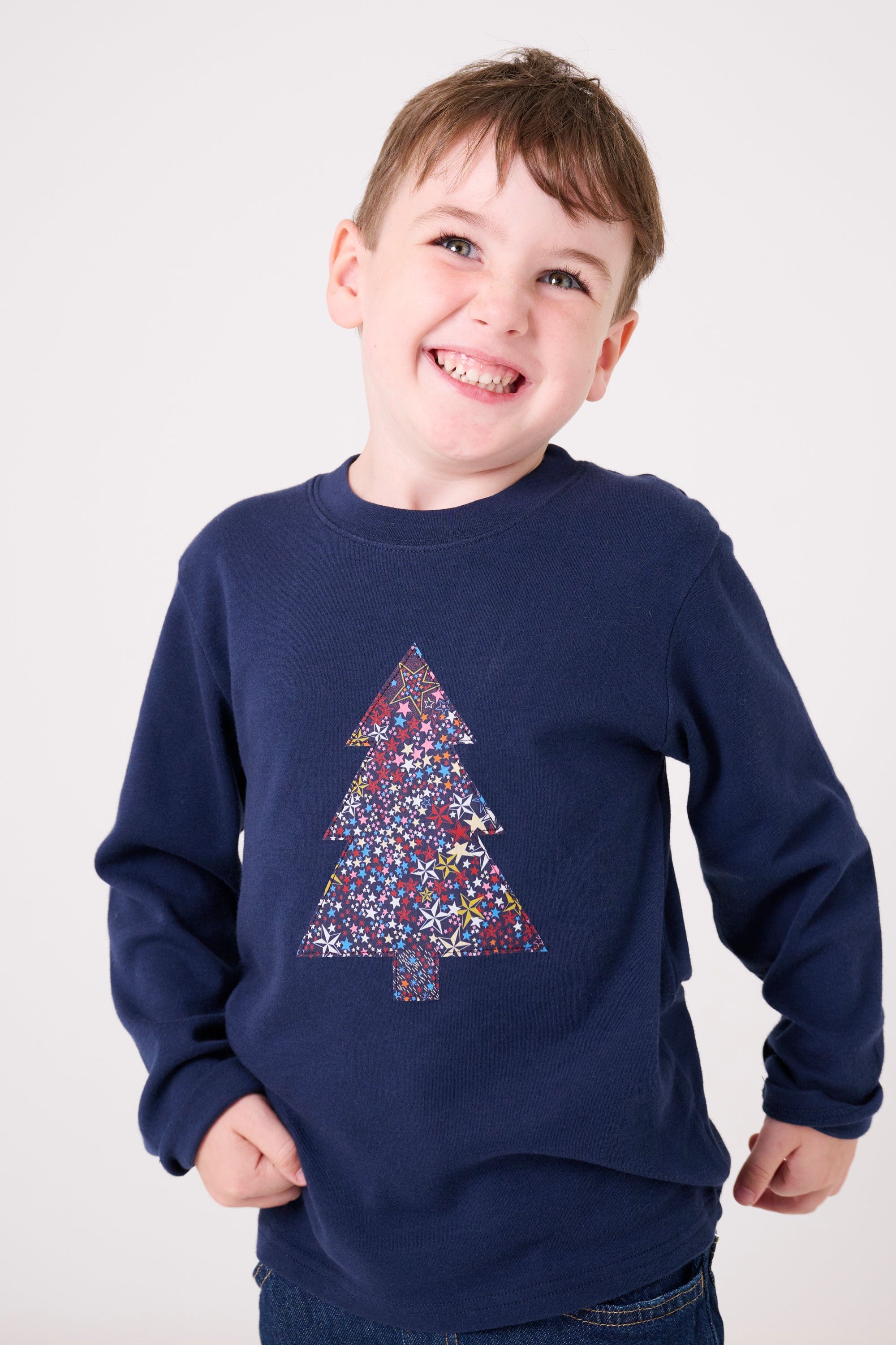 A 3 year old boy wearing a navy long sleeve Christmas t-shirt with a Christmas tree on the front made from Liberty Adelajda's Wish star print on the front.
