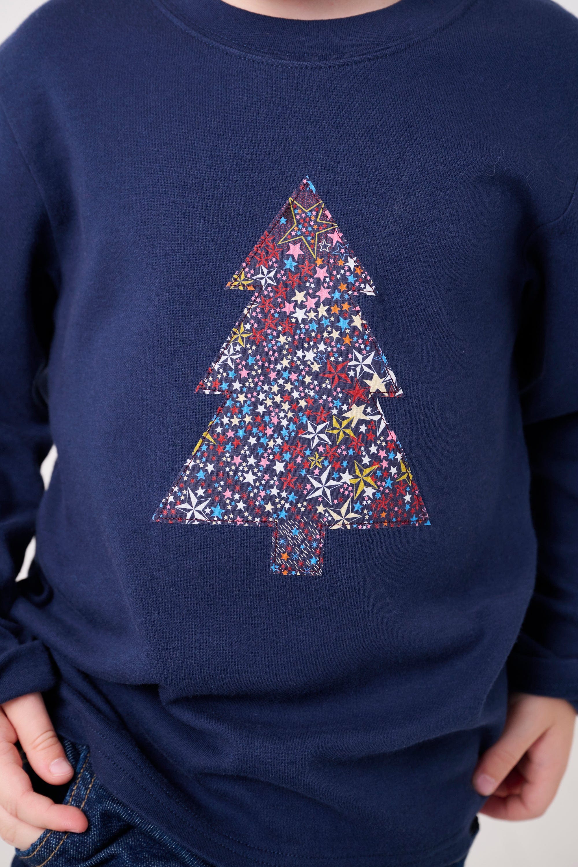 A 3 year old boy wearing a navy long sleeve Christmas t-shirt with a Christmas tree on the front made from Liberty Adelajda's Wish star print on the front