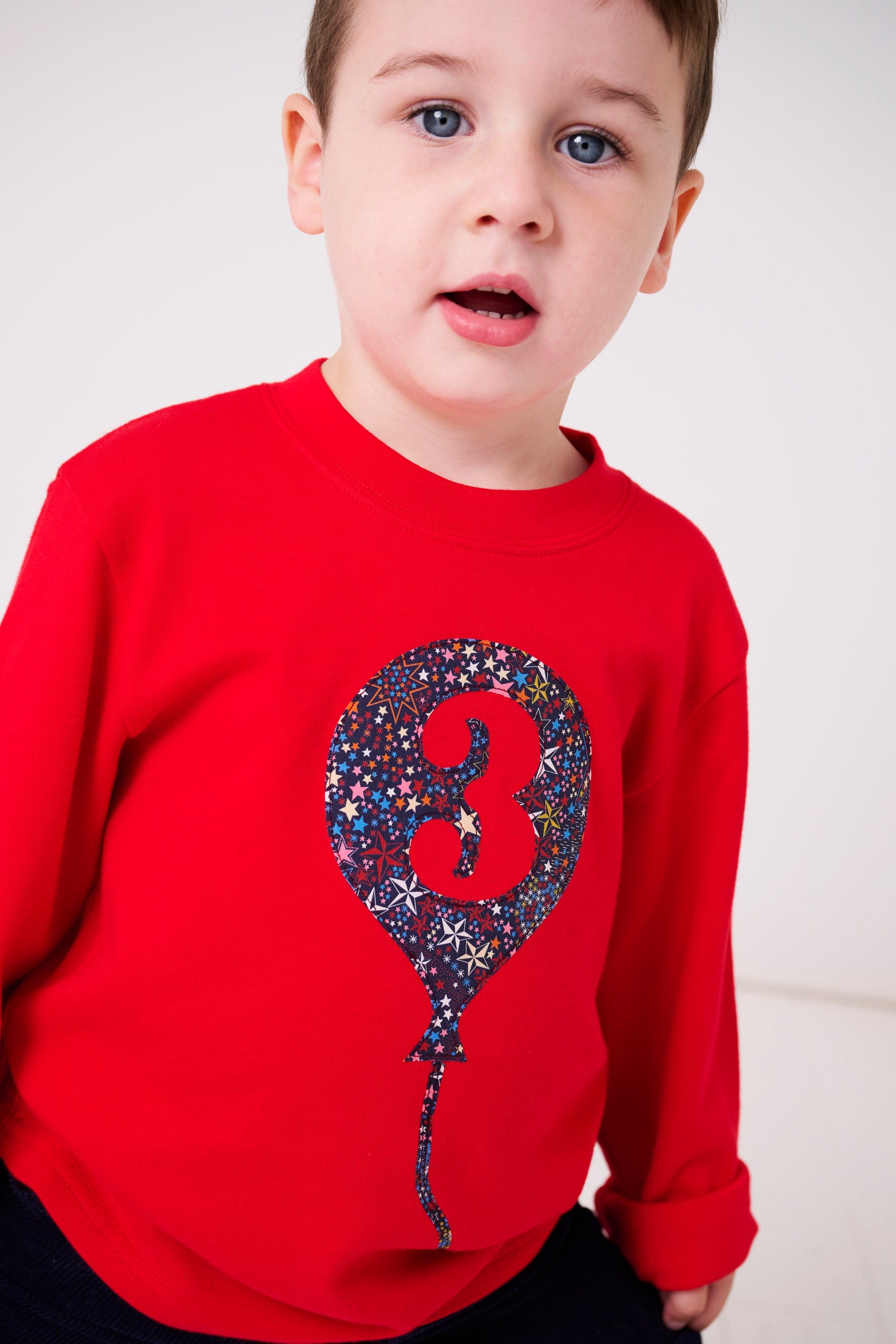 A 3 year old wearing a red cotton t-shirt with a liberty star print birthday balloon on the front with a  number 3 on it.