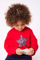 A 3 year old girl wearing a red Christmas t-shirt with a liberty floral Christmas star appliquéd on the front. She is holding a matching crown and wand.