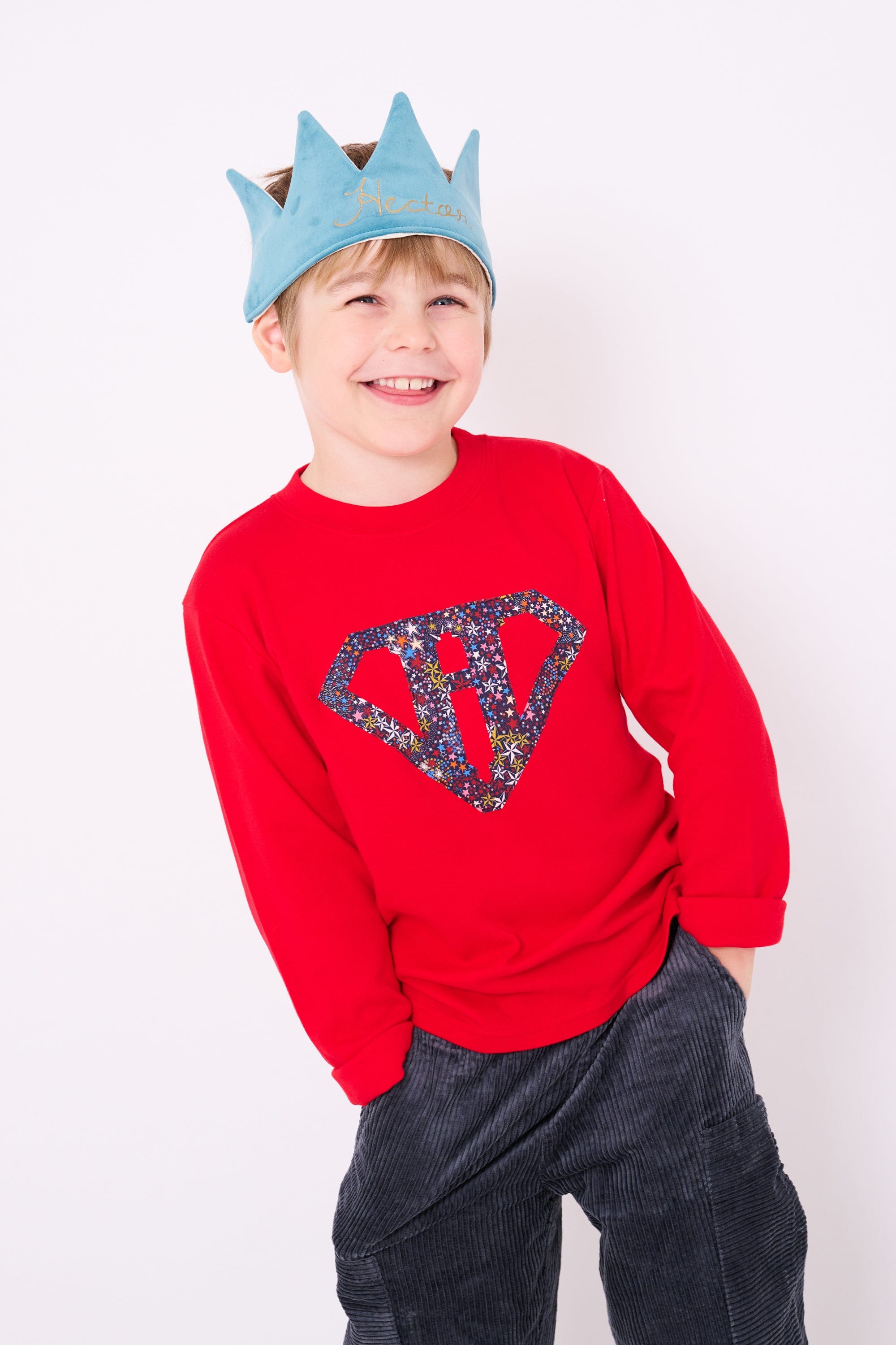 A smiling 5 year old boy wearing a festive red cotton personalised t-shirt with his initial in a superhero emblem Liberty star Adelajda's Wish fabric appliquéd on the front.