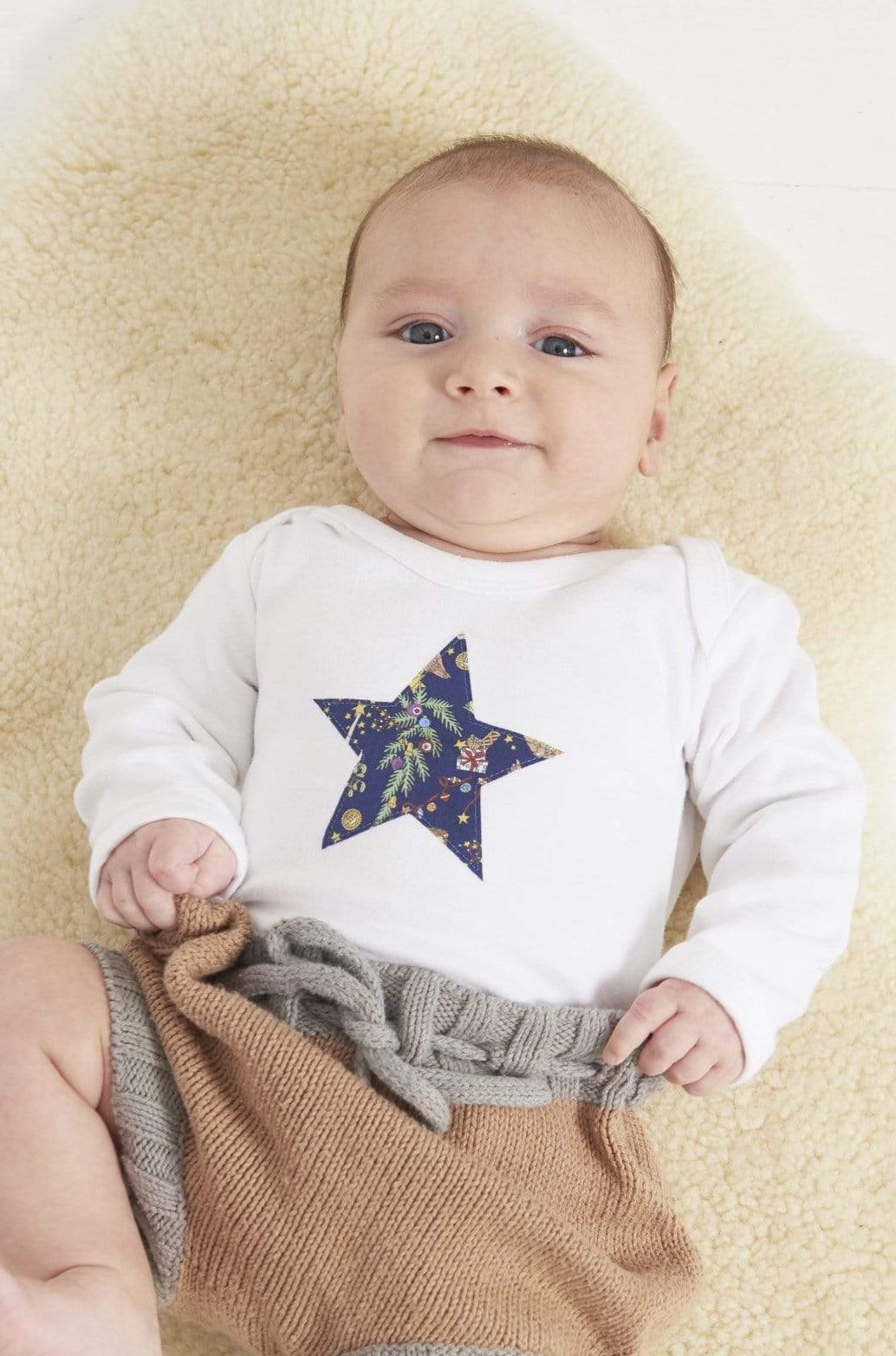 A baby wearing a white bodysuit babygrow with a Liberty Christmas print festive star sewn on the front.