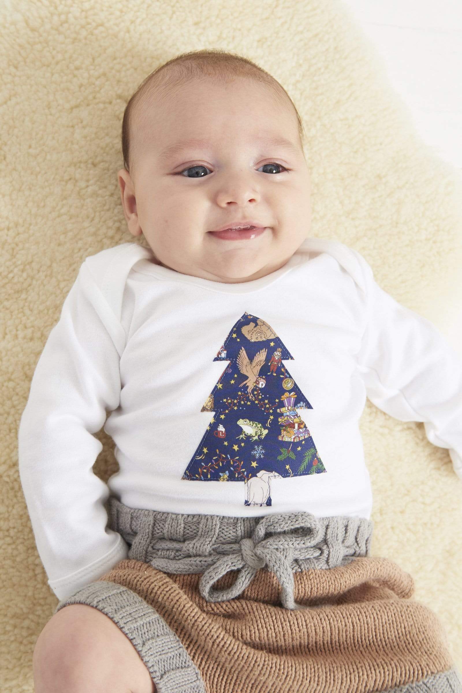 A baby wearing a white bodysuit babygrow with a Liberty Christmas print Christmas tree sewn on the front.