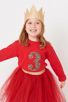 A 3 year old girl wearing a red 3rd birthday t-shirt with a liberty floral number 3 appliquéd on the front. 