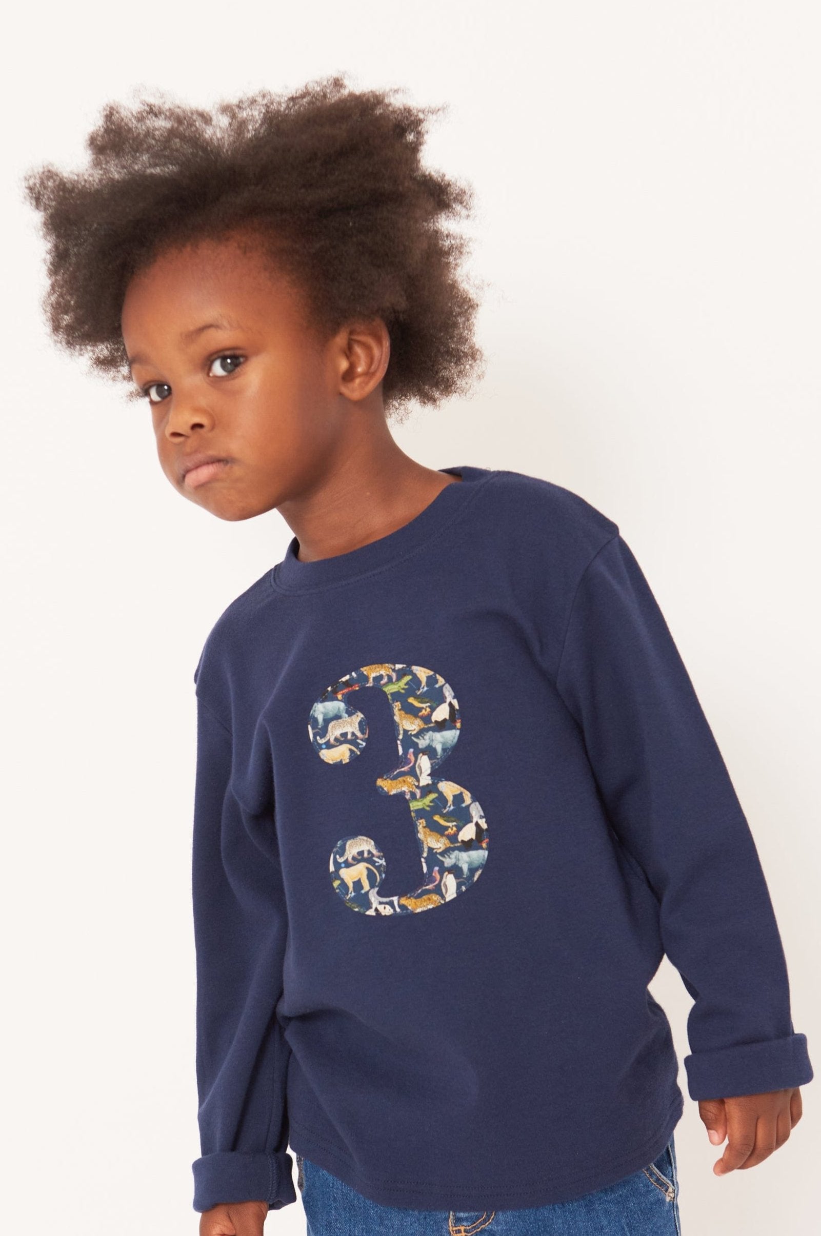 A 3 year old boy wearing a navy long sleeve t-shirt with a  number 3 on the front on in Liberty Quey Zoo animal print fabric.