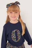 A smiling 5 year old girl wearing a personalised cotton navy long sleeve tee with her initial on the front made from Quey Zoo  animal Liberty print fabric.