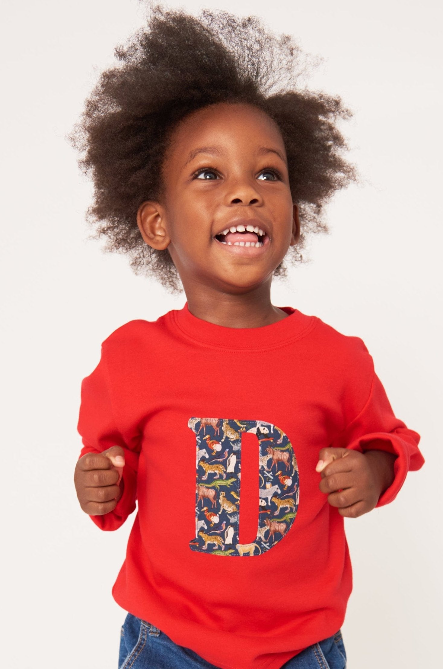 A smiling 2 year old boy wearing a personalised red t-shirt with his initial on the front made out of a Liberty zoo animal print Quey 2.