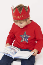 A 5 year old boy wearing a red Christmas t-shirt with a festive star on the front in a Liberty Christmas animals print. He is wearing a red velvet gown, reading a Christmas book.