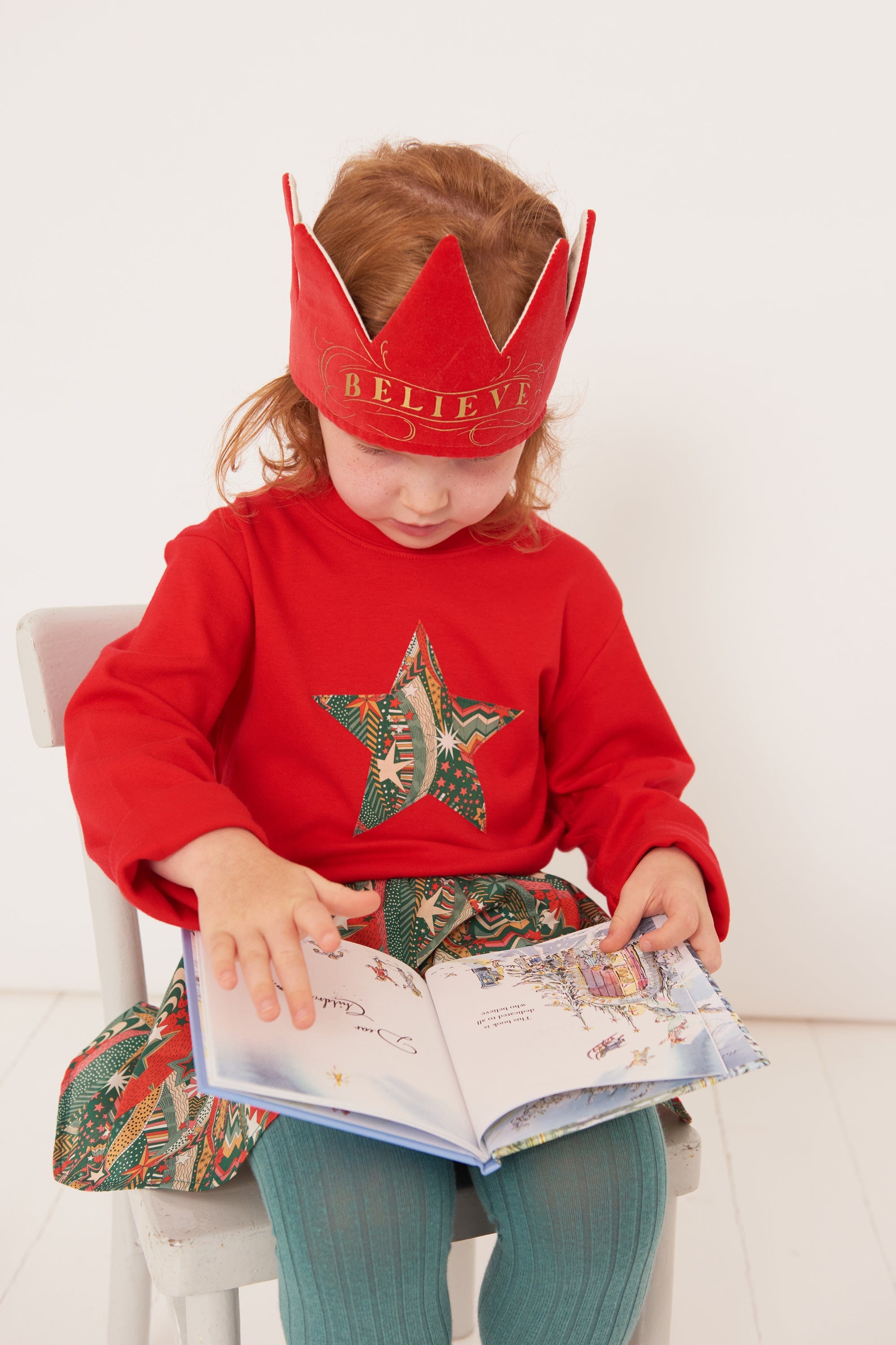 A 4 year old girl wearing a red Christmas t-shirt with a festive star on the front in a starry Liberty print, wearing a crown and reading a christmas book.